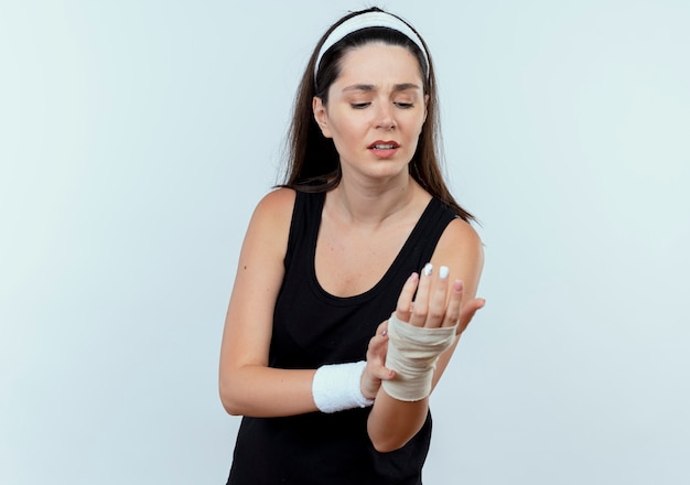 Joven mujer fitness en diadema mirando su muñeca vendada sintiendo dolor de pie sobre la pared blanca