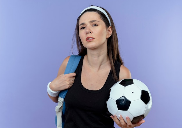 Joven mujer fitness en diadema con mochila sosteniendo un balón de fútbol mirando confundido de pie sobre la pared azul