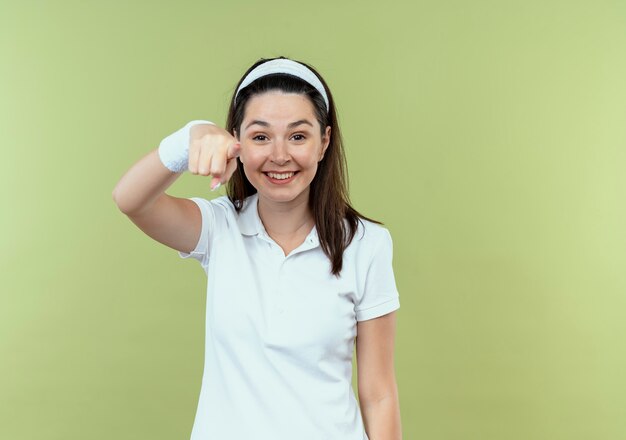 Joven mujer fitness en diadema sonriendo alegremente apuntando con el dedo de pie sobre la pared de luz