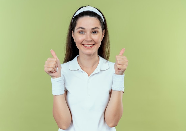 joven mujer fitness en diadema sonriendo alegremente mostrando los pulgares para arriba de pie sobre la pared de luz