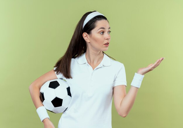 Joven mujer fitness en diadema sosteniendo un balón de fútbol presentando con el brazo de su mano mirando sorprendido de pie sobre la pared de luz