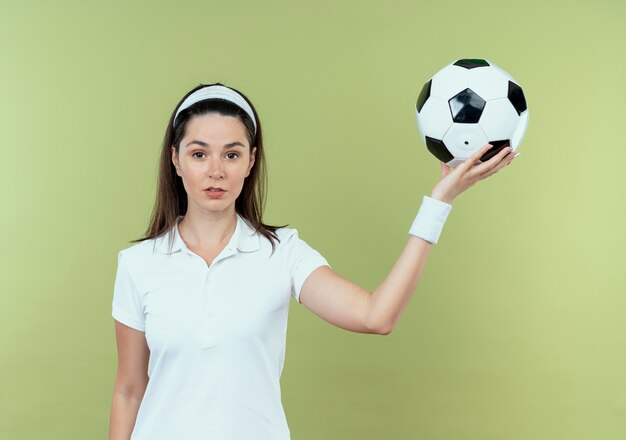 Joven mujer fitness en diadema sosteniendo un balón de fútbol con rostro serio de pie sobre la pared de luz