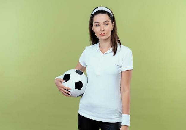 Joven mujer fitness en diadema sosteniendo un balón de fútbol con una seria expresión de confianza de pie sobre una pared de luz