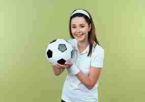 Foto gratuita joven mujer fitness en diadema sosteniendo un balón de fútbol sonriendo alegremente de pie sobre la pared de luz