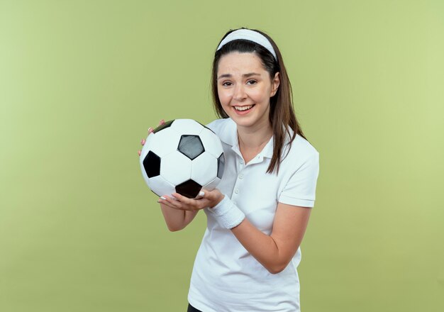 Joven mujer fitness en diadema sosteniendo un balón de fútbol sonriendo alegremente de pie sobre la pared de luz
