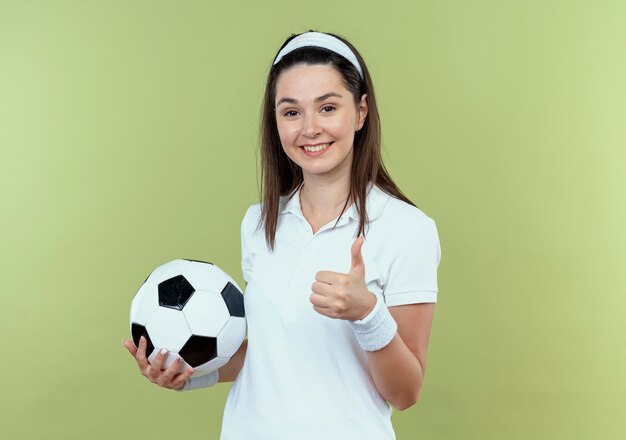 Joven mujer fitness en diadema sosteniendo un balón de fútbol sonriendo mostrando los pulgares para arriba de pie sobre la pared de luz