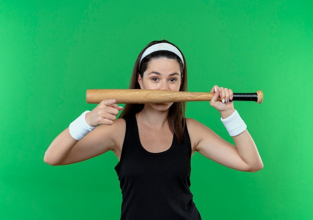 Joven mujer fitness en diadema sosteniendo un bate de béisbol apuntando con el dedo sonriendo de pie sobre la pared verde