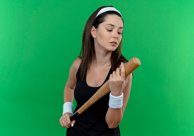 Joven mujer fitness en diadema sosteniendo un bate de béisbol mirándolo con cara seria de pie sobre la pared verde