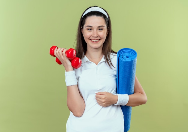 Joven mujer fitness en diadema sosteniendo dos mancuernas y estera de yoga sonriendo de pie sobre la pared de luz