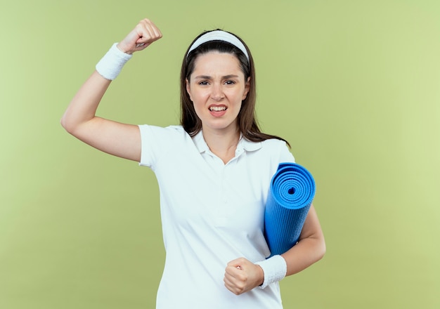 Joven mujer fitness en diadema sosteniendo estera de yoga levantando el puño con cara enojada de pie sobre la pared de luz