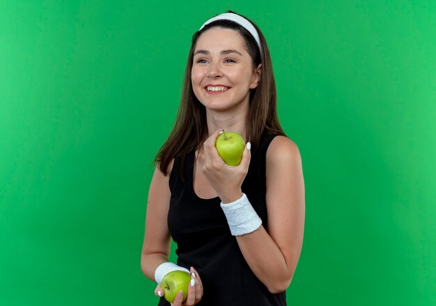 Joven mujer fitness en diadema sosteniendo manzanas verdes sonriendo con cara feliz de pie sobre la pared verde