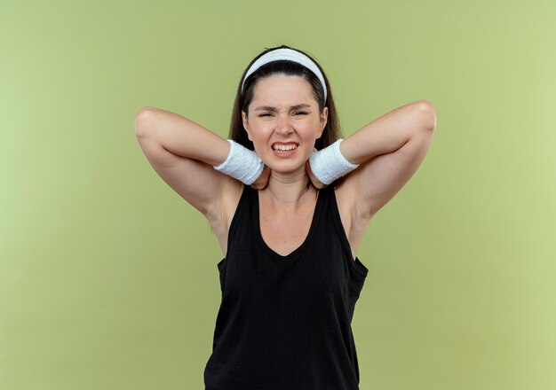 Joven mujer fitness en diadema tocando su cuello mirando malestar sintiendo dolor de pie sobre la pared de luz