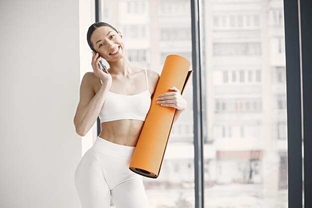 Joven mujer fitness lista para hacer ejercicio sosteniendo una alfombra de yoga naranja mientras usa un teléfono