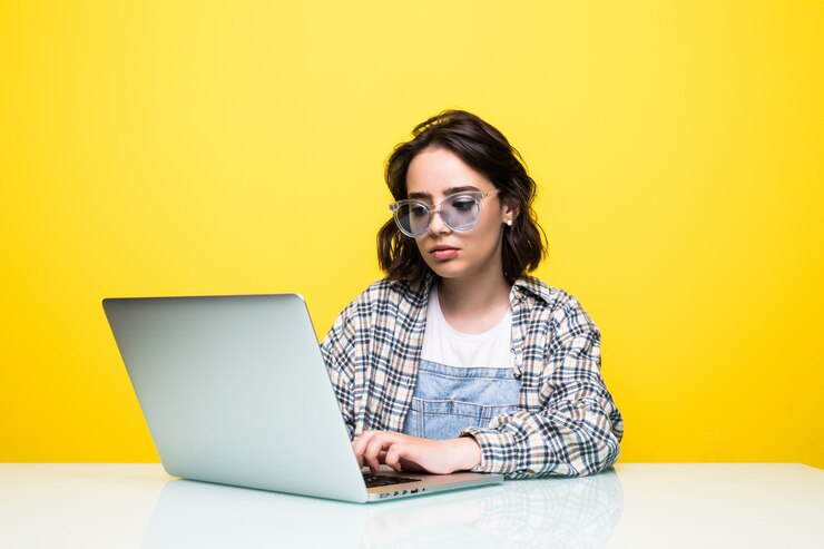 Foto gratuito joven mujer fresca positiva con gafas de sol usando laptop y sonriendo aislado sobre amarillo