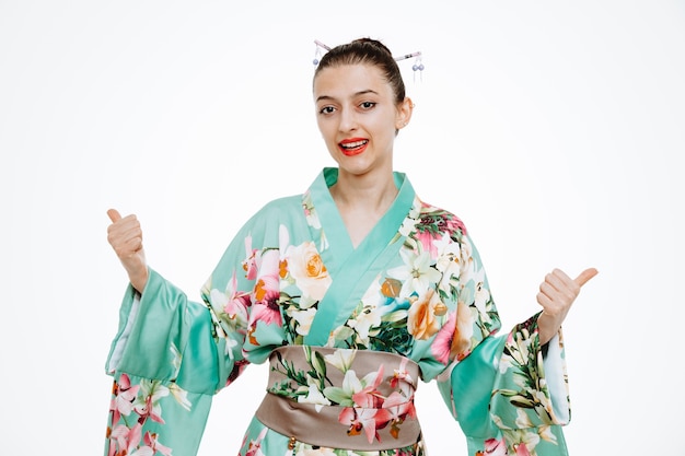 Joven mujer geisha en kimono tradicional japonés mirando al frente feliz y positivo mostrando los pulgares para arriba de pie sobre la pared blanca