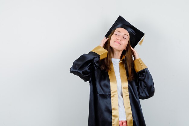 Joven mujer graduada cogidos de la mano en el cabello con traje académico y mirando esperanzado. vista frontal.