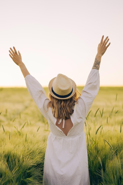 joven mujer hermosa con cabello largo rubio en un vestido blanco en un sombrero de paja recoge flores en un campo de trigo. Pelo volador al sol, verano. Hora de los soñadores, atardecer dorado.