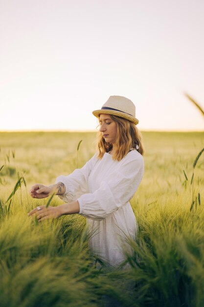 joven mujer hermosa con cabello largo rubio en un vestido blanco en un sombrero de paja recoge flores en un campo de trigo. Pelo volador al sol, verano. Hora de los soñadores, atardecer dorado.