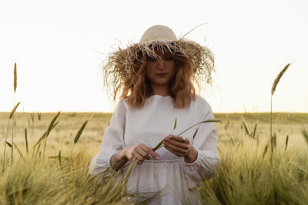 joven mujer hermosa con cabello largo rubio en un vestido blanco en un sombrero de paja recoge flores en un campo de trigo. Pelo volador al sol, verano. Hora de los soñadores, atardecer dorado.