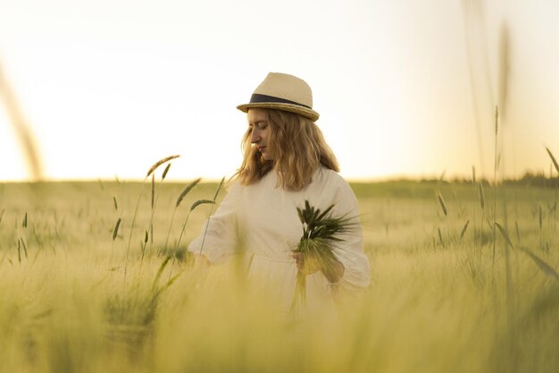 joven mujer hermosa con cabello largo rubio en un vestido blanco en un sombrero de paja recoge flores en un campo de trigo. Pelo volador al sol, verano. Hora de los soñadores, atardecer dorado.