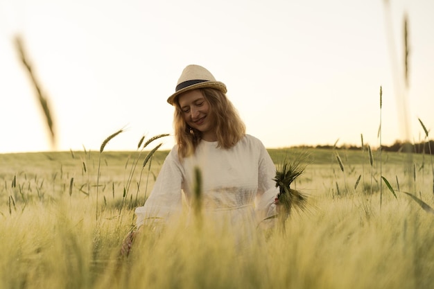 joven mujer hermosa con cabello largo rubio en un vestido blanco en un sombrero de paja recoge flores en un campo de trigo. Pelo volador al sol, verano. Hora de los soñadores, atardecer dorado.