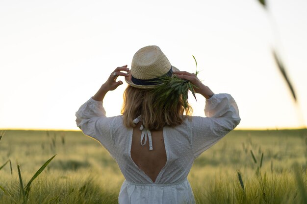 joven mujer hermosa con cabello largo rubio en un vestido blanco en un sombrero de paja recoge flores en un campo de trigo. Pelo volador al sol, verano. Hora de los soñadores, atardecer dorado.