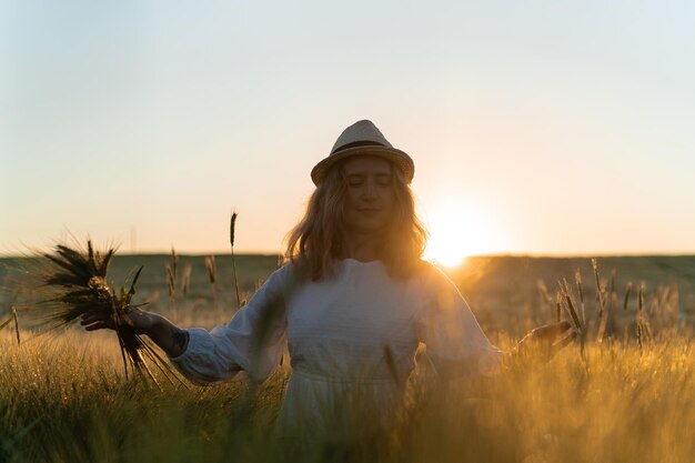 joven mujer hermosa con cabello largo rubio en un vestido blanco en un sombrero de paja recoge flores en un campo de trigo. Pelo volador al sol, verano. Hora de los soñadores, atardecer dorado.