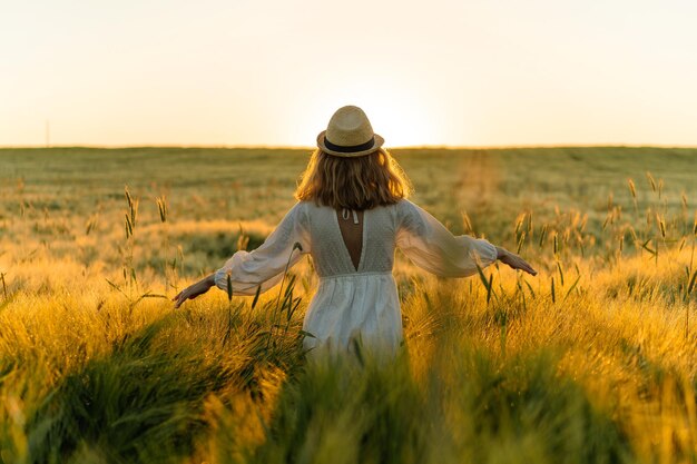 joven mujer hermosa con cabello largo rubio en un vestido blanco en un sombrero de paja recoge flores en un campo de trigo. Pelo volador al sol, verano. Hora de los soñadores, atardecer dorado.