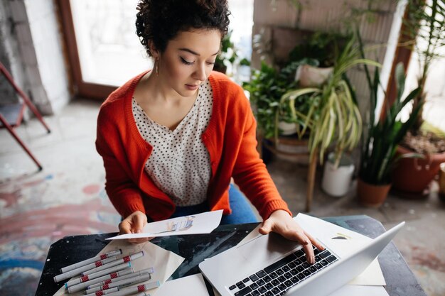 Joven mujer hermosa con cabello rizado oscuro sentada en la mesa trabajando cuidadosamente en una laptop con ilustración de moda en la mano mientras pasa tiempo en un taller moderno y acogedor con grandes ventanas