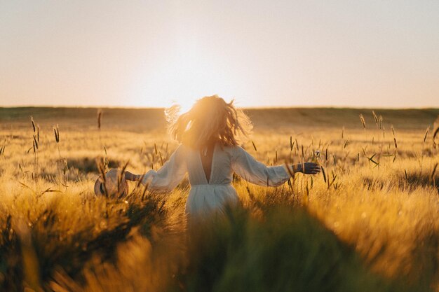 joven mujer hermosa con el pelo largo y rubio con un vestido blanco en un campo de trigo temprano en la mañana al amanecer. El verano es el momento de los soñadores, el pelo volador, una mujer corriendo por el campo bajo los rayos.