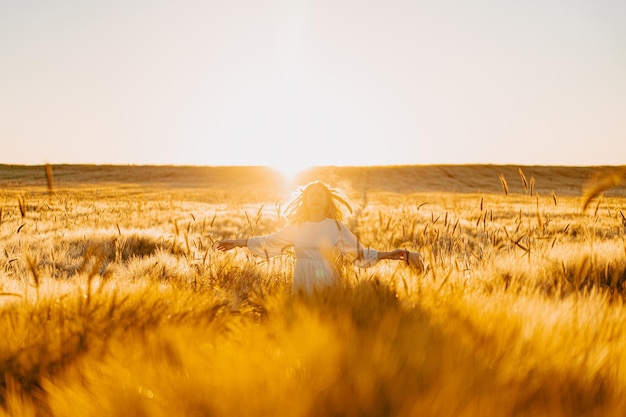 joven mujer hermosa con el pelo largo y rubio con un vestido blanco en un campo de trigo temprano en la mañana al amanecer. El verano es el momento de los soñadores, el pelo volador, una mujer corriendo por el campo bajo los rayos