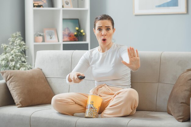 joven mujer hermosa con ropa informal sentada en un sofá en el interior de casa con un cubo de palomitas de maíz sosteniendo un mando a distancia mirando la cámara preocupada haciendo un gesto de parada con la mano