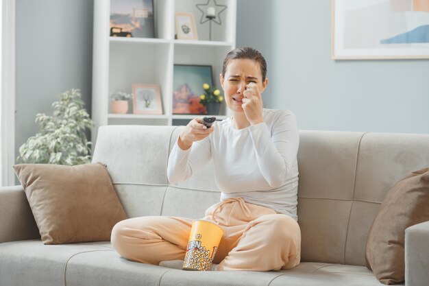 Joven mujer hermosa con ropa informal sentada en un sofá en el interior de su casa con un cubo de palomitas de maíz sosteniendo un drama de observación remota llorando molesto y triste frotándose los ojos pasando el fin de semana en casa