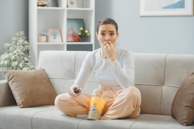 joven mujer hermosa con ropa informal sentada en un sofá en el interior de su casa con un cubo de palomitas de maíz sosteniendo un mando a distancia mirando la cámara asustada y nerviosa mordiéndose las uñas