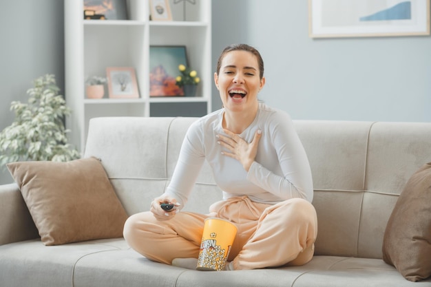 joven mujer hermosa con ropa informal sentada en un sofá en el interior de su casa con un cubo de palomitas de maíz sosteniendo un mando a distancia mirando a la cámara feliz y alegre sonriendo ampliamente pasando tiempo en casa