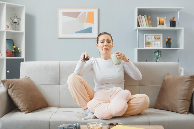 joven mujer hermosa con ropa informal sentada en un sofá en el interior de su casa sosteniendo un mando a distancia y una taza de té con aspecto confundido y disgustado mientras ve la televisión pasando el fin de semana en casa
