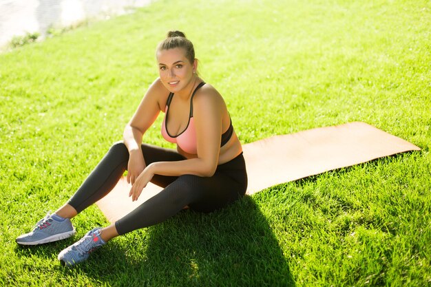 Joven mujer hermosa de talla grande con top deportivo y polainas sentada en una alfombra de yoga mirando felizmente a la cámara mientras pasa tiempo en la hierba verde en el parque