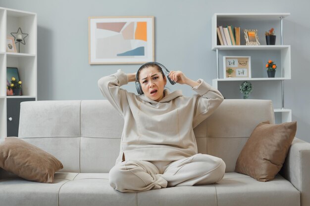 Joven mujer hermosa vestida de casa con auriculares sentada en un sofá en el interior de casa mirando a la cámara confundida y disgustada