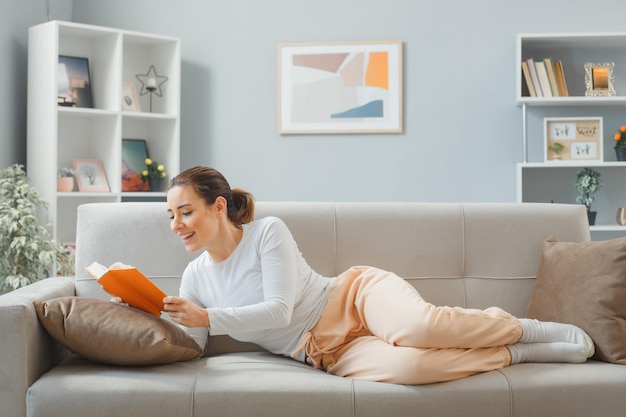 Joven mujer hermosa vestida de casa relajándose en un sofá en el interior de su casa leyendo un libro feliz y sonriendo positivamente pasando el fin de semana en casa