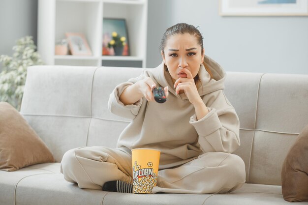 joven mujer hermosa vestida de casa sentada en un sofá en el interior de la casa con un cubo de palomitas de maíz sosteniendo a distancia viendo la televisión confundida y disgustada