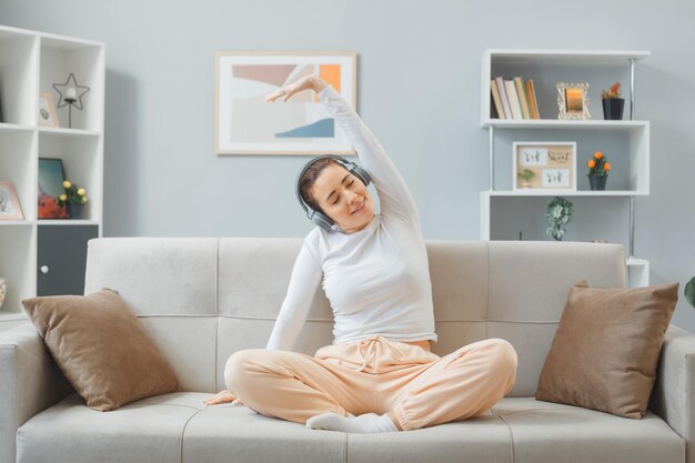 Joven mujer hermosa vestida de casa sentada en un sofá en el interior de su casa con auriculares felices y positivos relajándose estirándose disfrutando de su tiempo pasando el fin de semana en casa