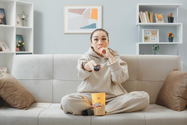Joven mujer hermosa vestida de casa sentada en un sofá en el interior de su casa con un cubo de palomitas de maíz sosteniendo un control remoto viendo la televisión preocupada y nerviosa pasando tiempo en casa