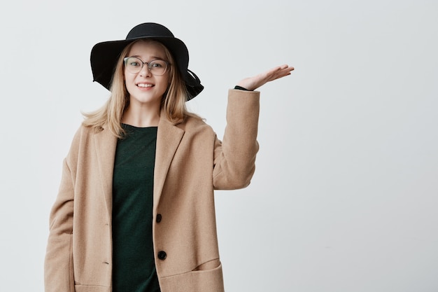 Foto gratuita joven mujer infeliz, gesticulando con las palmas abiertas, mirando hacia arriba, harta, quiere salir pero tiene miedo a la lluvia. estudiante con abrigo y sombrero confundida por las malas condiciones climáticas.