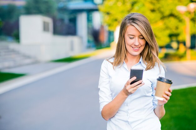 Joven mujer inteligente de lectura profesional con teléfono. Mujer de negocios de lectura de noticias o mensajes de texto sms en smartphone, mientras que el consumo de café en el descanso del trabajo.