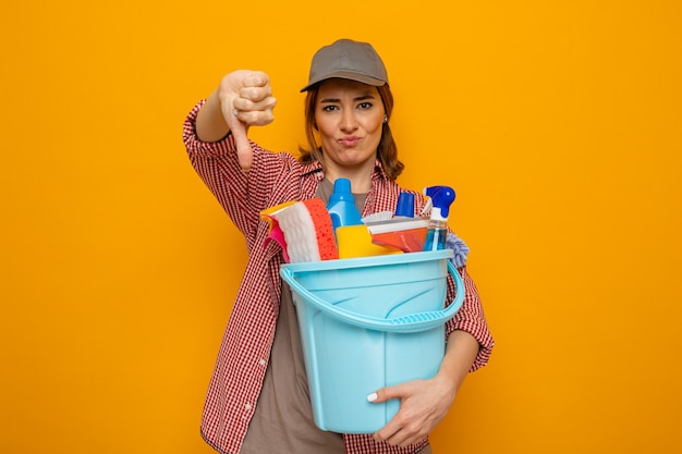 Joven mujer de limpieza en camisa a cuadros y gorra sosteniendo un balde con herramientas de limpieza mirando a la cámara disgustado mostrando los pulgares hacia abajo de pie sobre fondo naranja
