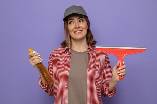 Joven mujer de limpieza en camisa a cuadros y gorra sosteniendo una botella de productos de limpieza y un trapeador mirando hacia arriba sonriendo alegremente de pie sobre fondo púrpura