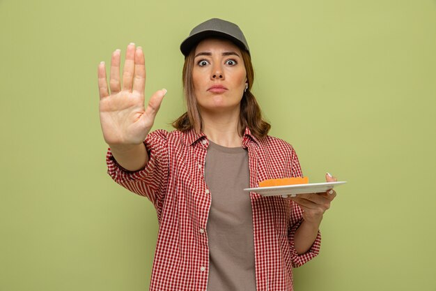 Joven mujer de limpieza en camisa a cuadros y gorra sosteniendo la placa y la esponja mirando a la cámara con cara seria haciendo gesto de parada con la mano de pie sobre fondo verde