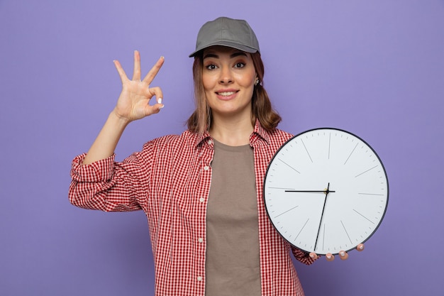 Foto gratuita joven mujer de limpieza en camisa a cuadros y gorra sosteniendo reloj mirando haciendo bien firmar sonriendo alegremente