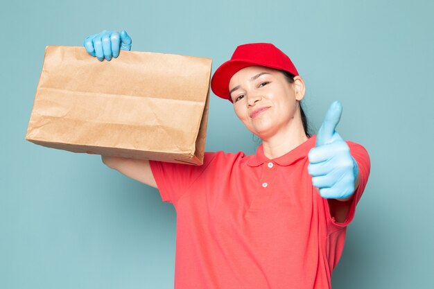 Joven mujer mensajero en camiseta rosa gorra roja guantes azules caja en la pared azul