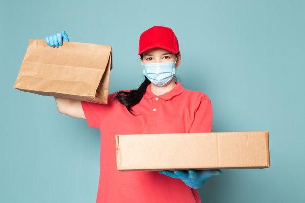 Joven mujer mensajero en camiseta rosada gorra roja máscara azul estéril guantes azules con caja en la pared azul